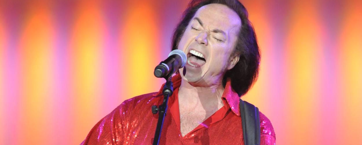 The image captures a close-up of a male singer on stage, engrossed in his performance. He is wearing a striking, bright red, sequined shirt that shimmers under the stage lights. The shirt has a collar and is unbuttoned slightly at the neck, revealing a patch of chest hair. His face is contorted in a passionate expression as he sings, with his eyes closed tightly and his mouth wide open, revealing his teeth and tongue. He has dark, slicked-back hair that frames his face, and his skin appears to have some tex