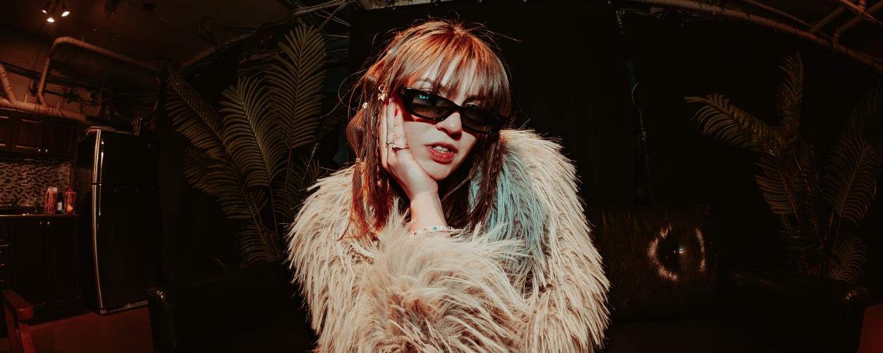 The image is a medium shot of a young woman posing in a dimly lit, somewhat cluttered indoor space. The woman is in the center of the frame, looking directly at the camera. She has reddish-brown hair with bangs and is wearing large, dark sunglasses with a thick black frame. Her left hand is resting on her cheek, with her fingers gently touching her face. She is wearing a light-colored, fluffy, faux fur jacket that covers most of her upper body. Several rings are visible on her left hand, and a beaded bracel