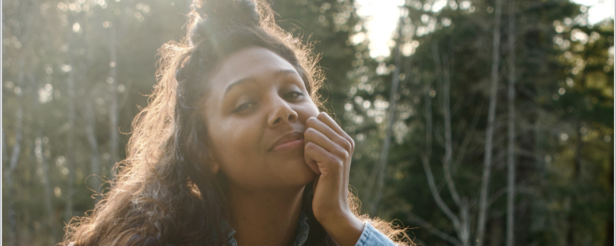 The image is a medium shot portrait of a young woman with dark, curly hair, some of which is pulled up into a messy bun at the top of her head. She is looking towards the camera with a gentle, slightly upward gaze and a soft smile playing on her lips. Her right hand is brought up to her face, with her fingers resting lightly against her cheek and chin, a pose that suggests contemplation or contentment.