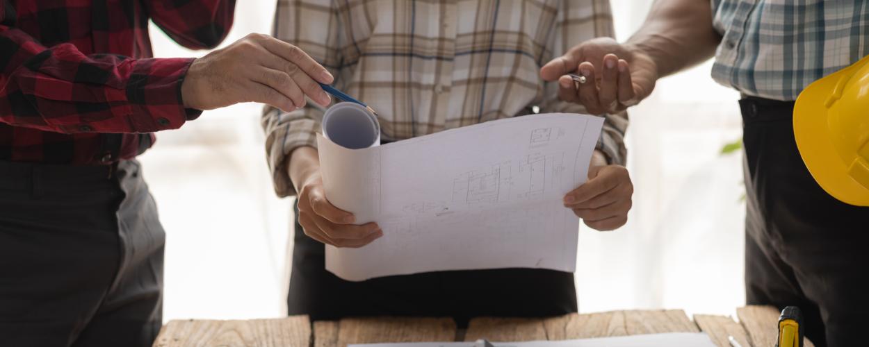 Three people stand over architectural plans and a model home on top of some paperwork.