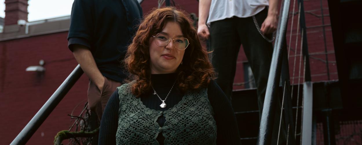 The image is an outdoor, medium shot of three people standing on a metal staircase against a brick building. The central figure is a young woman with curly, reddish-brown hair, wearing glasses with thin, gold-toned frames. She has fair skin and is looking directly at the camera with a slight smile. She is wearing a dark turtleneck shirt and a distinctive, dark green crocheted vest with a floral pattern and buttons down the front. A delicate silver necklace with a locket-shaped pendant hangs around her neck