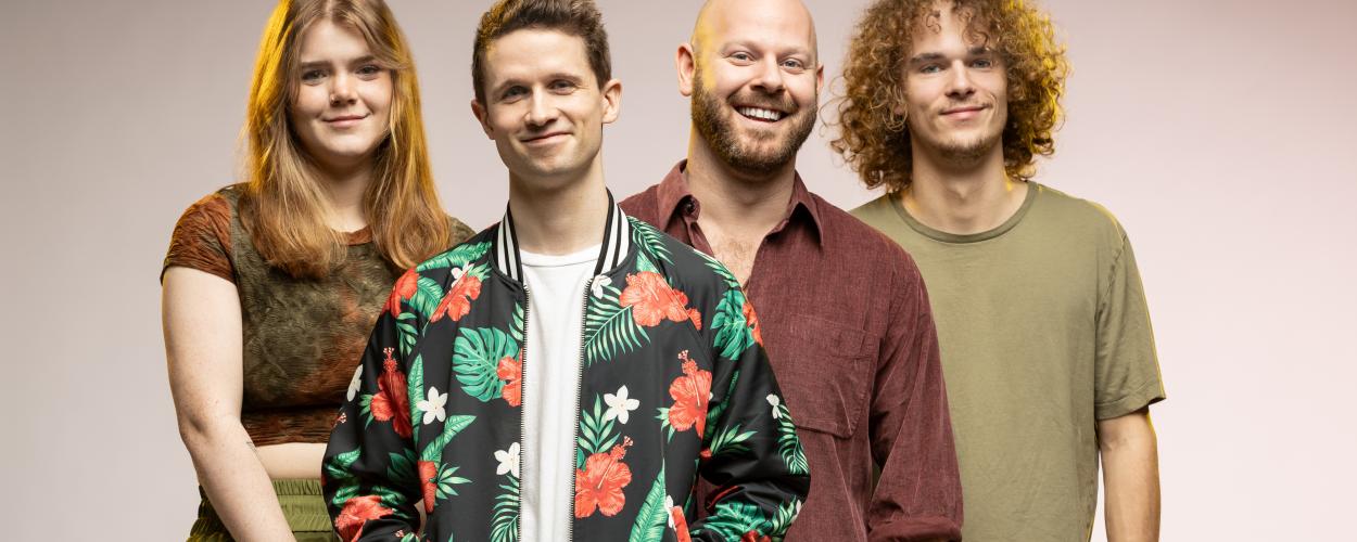 A group of four young adults, two women and two men, stand in a line facing forward against a neutral, light grey to pink gradient background. The lighting is warm and falls primarily on the faces and upper bodies of the subjects, creating soft shadows.