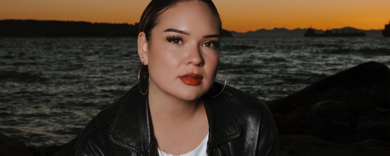 The image is a medium shot portrait of a young woman seated outdoors by the water, likely at dusk or dawn given the warm tones of the sky. The woman is positioned slightly off-center to the right, with her gaze directed straight at the camera. She has her hair pulled back neatly, revealing her face and ears. She is wearing a black leather jacket over a white t-shirt. Her makeup is noticeable, with emphasis on her eyes, which are adorned with false eyelashes, and her lips, painted in a bold reddish-orange hu