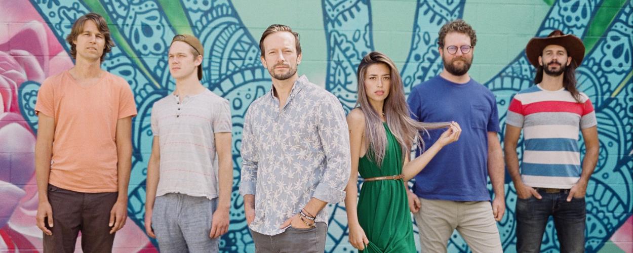 The image shows a group of six people standing side-by-side in front of a vibrant, colorful mural. The mural features intricate blue and green patterns that resemble mandala or floral designs, adding a striking artistic backdrop.