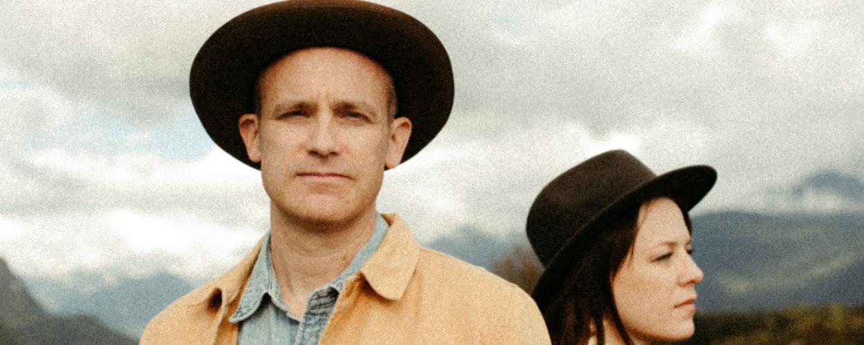 The image features a medium shot of two people, a man and a woman, positioned in the foreground against a blurred outdoor landscape.  The man is on the left side of the frame, facing the camera directly. He is bald and has a serious expression. He is wearing a wide-brimmed brown hat, a light blue denim shirt, and an open, mustard-yellow jacket. His jacket has a soft, textured appearance.