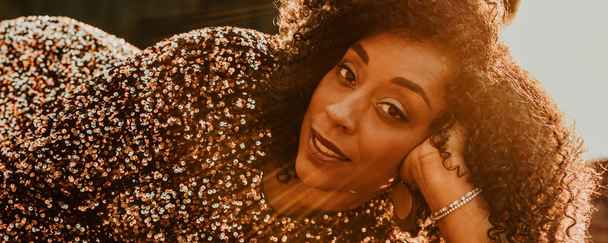 The image is a close-up, portrait-style photograph of a Black woman with curly dark brown hair, looking towards the camera. She is lying down, with her head resting on her left hand. The woman is wearing a shimmering dress covered in small, iridescent sequins that appear to be shades of bronze, gold, and copper, creating a textured and eye-catching garment. The sequins reflect the warm, golden light, adding to the overall glow of the image