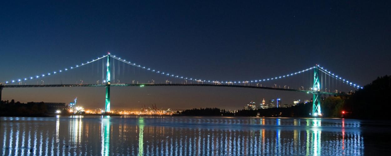 The Lions Gate Bridge seen at night with lights along the suspension cables. The water below reflect the bridges lights. 