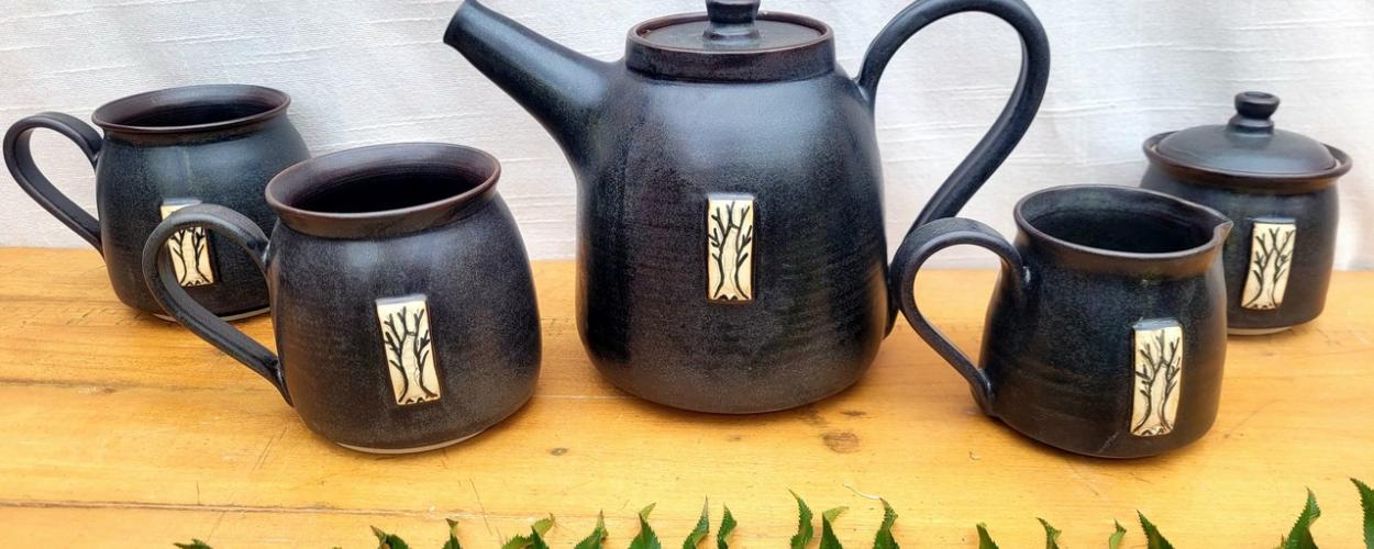 A rustic, handmade ceramic tea set is displayed on a wooden surface. The set includes a teapot, three mugs, and a lidded sugar bowl or creamer. All the pieces are a deep, matte black with a slight speckled texture, suggesting a natural clay body fired at a high temperature. Each item is adorned with a decorative rectangular plaque featuring a light-colored, stylized tree with bare branches. The interior rim of each mug and the teapot's lid and opening show a contrasting deep reddish-brown glaze.