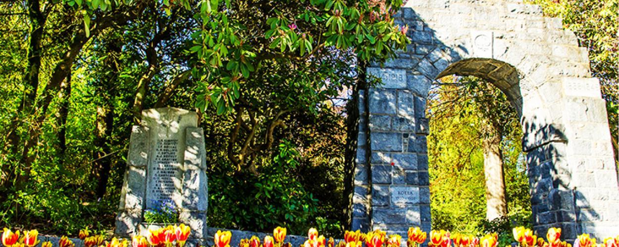 A stone arch and plaque sit at the edge of a forest surrounded by tulips.