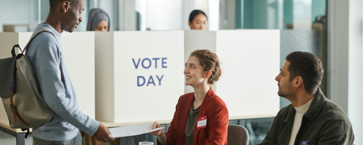 Two people wearing nametags sit at a table. One person is handing papers to someone standing in front of them. A sign in the background reads "Vote Day."