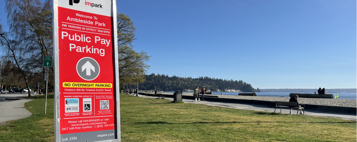 A photo of Ambleside beach on a clear blue-sky day. In the foreground is a red Public Pay Parking sign.
