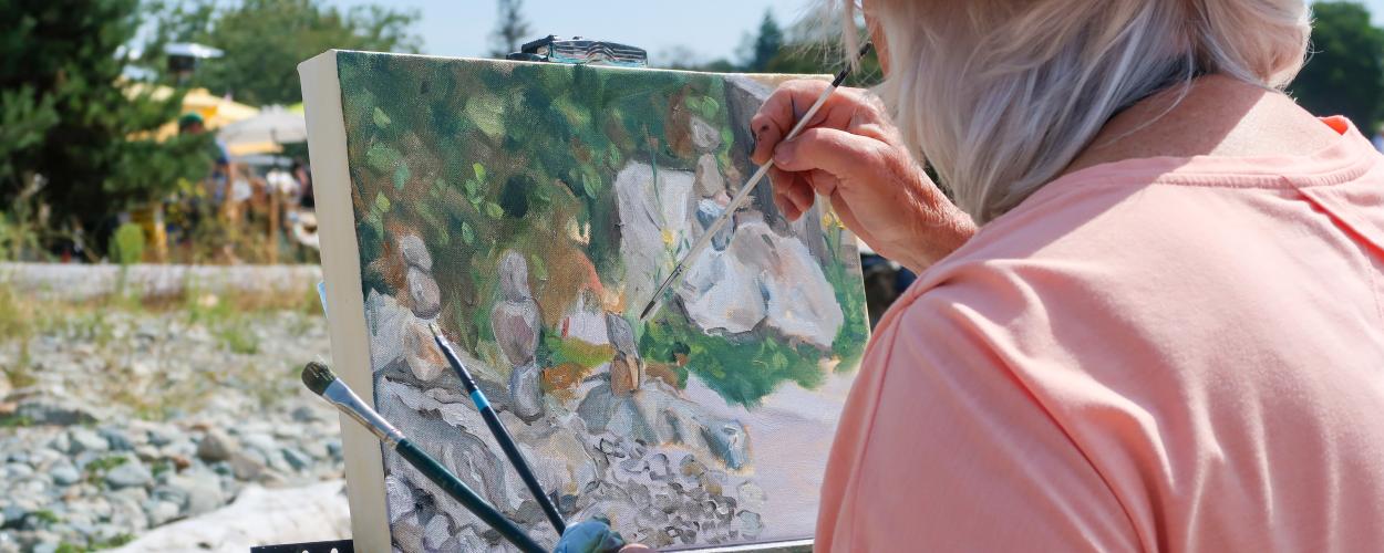 In a close-up, over-the-shoulder shot, an elderly person with white hair is painting outdoors. They are wearing a straw hat that casts a slight shadow over their face, and a salmon-pink t-shirt. The artist holds a thin paintbrush in their right hand, their fingers stained with paint, and is actively working on a canvas set on a dark easel. 