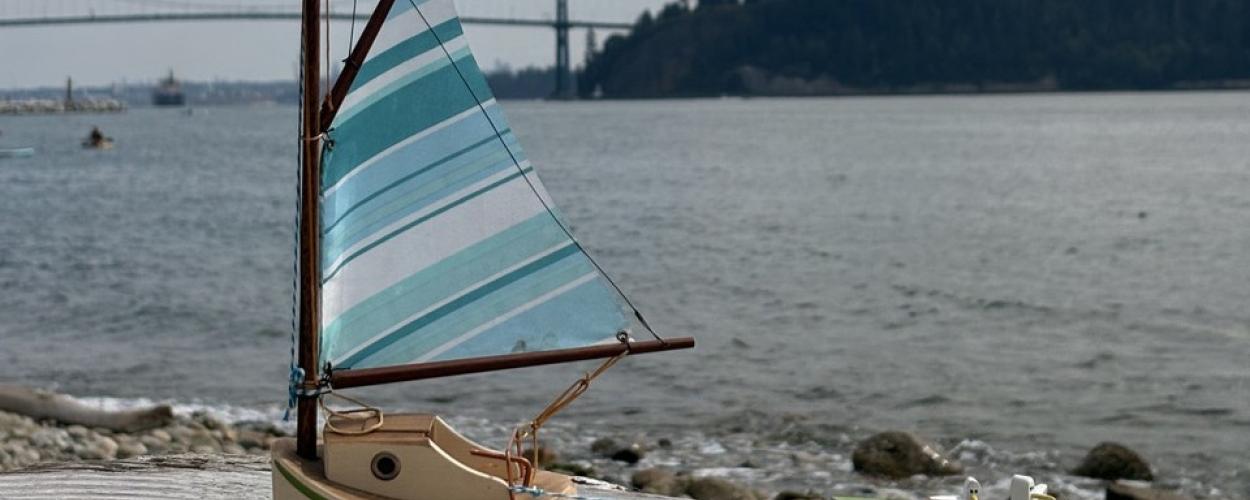 In the foreground of the image, two miniature toy boats are positioned on a weathered wooden surface that appears to be a pier or dock. The larger boat, on the left, is a sailboat with a predominantly light tan hull. It features a dark reddish-brown base and a thin green stripe just above it. The cabin area of the sailboat is a lighter tan color with a circular dark detail on its side. Its mast is made of wood, and the sail is the most striking element, displaying a pattern of alternating stripes in various