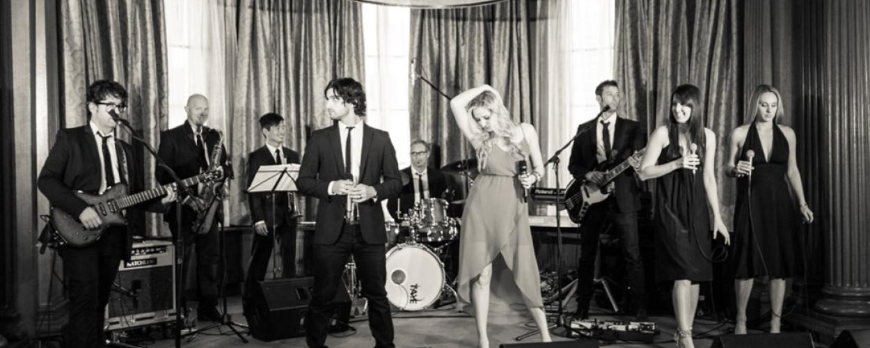A group of people is performing on stage, showcasing a musical performance. They are dressed in various clothing styles, including suits, while playing instruments such as a guitar and drums. The setting appears to be an indoor concert with a curtain backdrop. The image has a black and white aesthetic, adding to its artistic flair.