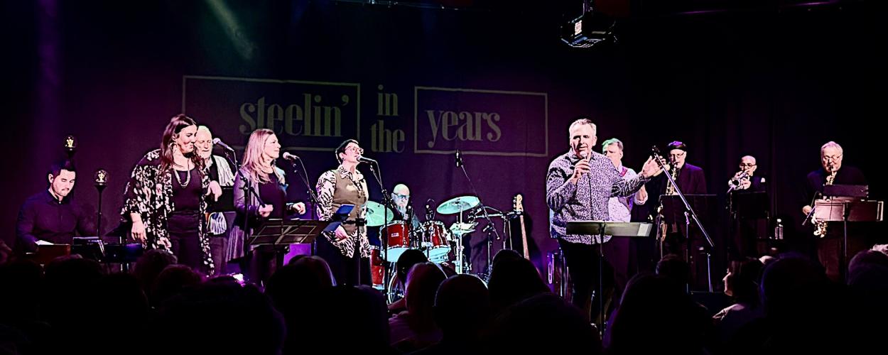 The image captures a live musical performance taking place on a stage. The background features a dark curtain with a large, illuminated banner that reads "steelin' in the years" in a stylized, outlined font. The banner has a purple hue, contrasting with the stage lighting.