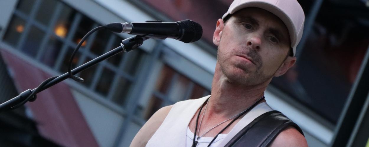 A male musician is performing on stage, holding an electric guitar and singing into a microphone. He is wearing a white tank top, a light pink baseball cap, and two silver necklaces with pendants. The guitar is a light brown color with a dark fretboard. The microphone is positioned in front of his face. The background is blurred but appears to be an outdoor venue with some structures and lights. The overall lighting suggests it is either daytime or early evening. The composition is a medium shot focusing on