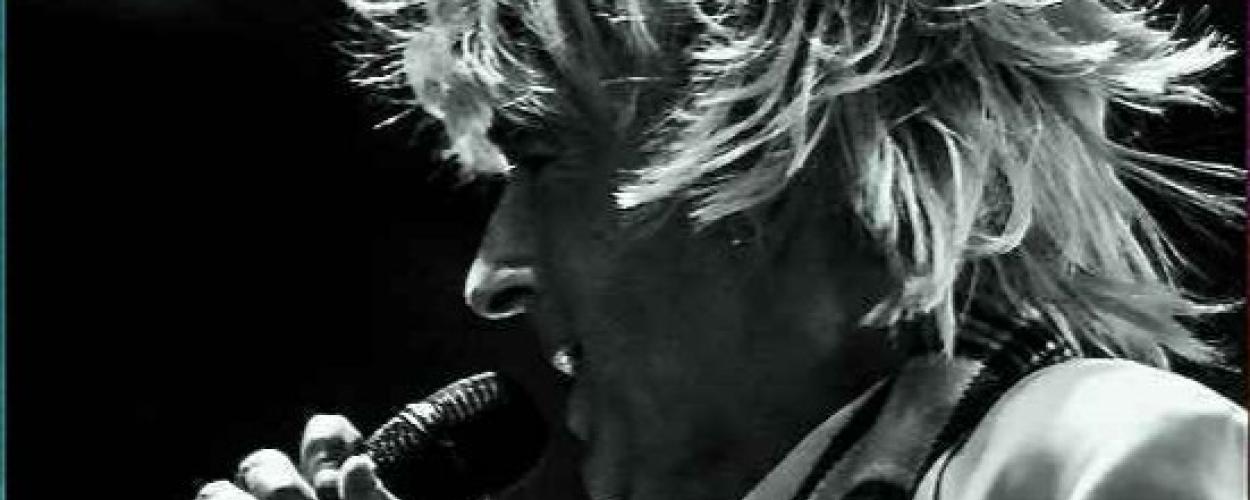 This is a black and white, close-up, profile shot of a male singer performing on stage. He has a full head of wild, spiky, blonde hair that frames his face and extends outward, suggesting movement. His head is tilted slightly downwards and to the left as he sings into a microphone held in his right hand. The microphone is dark and has a ribbed texture on its head. His mouth is open, and his eyes are not visible due to the angle.