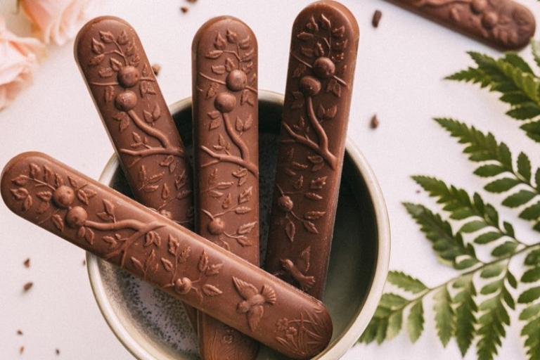 The image features a bowl filled with chocolate sticks, showcasing a delightful and visually appealing treat. The chocolate sticks likely have a smooth texture and rich flavor, making them an enticing snack or dessert option. The presence of the plant tag may suggest that the setting includes natural elements that enhance the overall aesthetic.