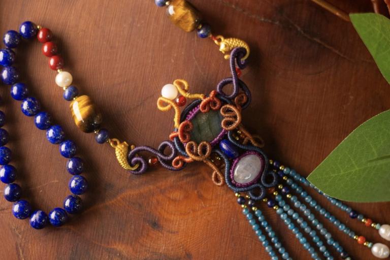The image displays a detailed and ornate necklace, intricately crafted with a variety of beads and macrame elements, laid out on a dark, textured wooden surface. The necklace is positioned diagonally across the frame, with a significant portion visible from the upper left to the lower right.