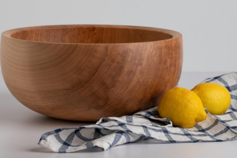 The image is a still life featuring a large, light-colored wooden bowl, two bright yellow lemons, and a blue and white checkered cloth. The setting appears to be a bright, minimally decorated studio with a white or light gray background and a white surface for the objects.