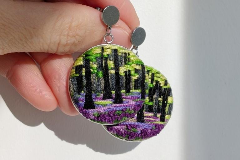 The image showcases a pair of handcrafted circular earrings designed with a nature-inspired embroidered scene. The earrings are being held by a person's hand, with the fingers gently supporting the lower edge of the earrings. The background is a plain, light-colored surface, likely a wall or table, with subtle shadows cast by the hand and earrings, suggesting a bright light source from the right.