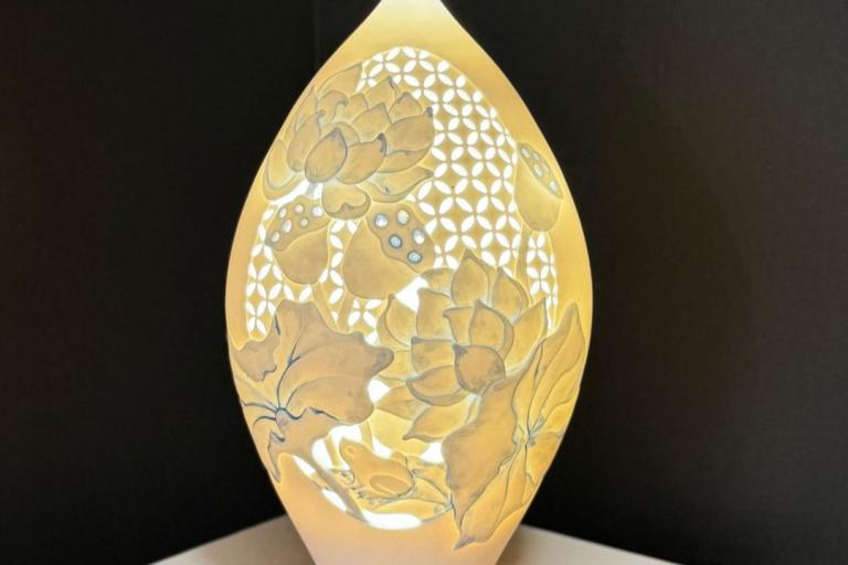 The image displays a beautifully illuminated porcelain vase with an intricate design. The vase has an elongated, slightly tapered shape, resembling a seed pod or a large teardrop, with a rounded bottom that transitions into a narrower top. It is predominantly rendered in warm, translucent tones of ivory and soft yellow, suggesting that it is lit from within, casting a gentle glow.