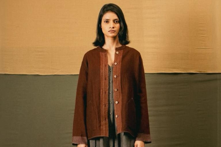 A young woman with shoulder-length dark hair and a serious expression stands facing the camera. She is positioned centrally in the frame, with her body from the chest up fully visible. She is wearing a dark reddish-brown, collarless jacket with a plaid lining visible underneath, the buttons down the front of the jacket are light brown and circular. The jacket has long sleeves that extend to her wrists, and two subtle pockets are visible on the front. Underneath the jacket, a dress or top with a black and wh