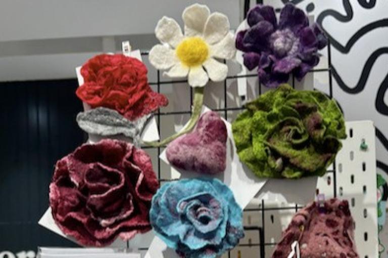 The image displays a collection of handcrafted felt flowers of various colors and designs, arranged on a black pegboard grid. The lighting appears to be artificial and slightly warm, illuminating the textured surfaces of the felt.