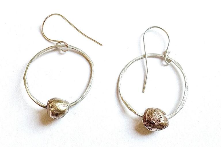 The image displays a pair of handcrafted hoop earrings positioned against a plain white background. Each earring consists of a circular hoop made of a silver-toned metal, with a slightly irregular and textured surface. A single, irregularly shaped bead or charm is suspended from the bottom of each hoop. These beads have a rough, organic texture and appear to be a darker, possibly bronze or oxidized silver, color, contrasting with the lighter hoops. One earring is slightly larger and positioned to the left, 