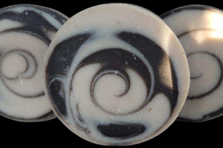 Three circular soaps are arranged in a close-up, shallow depth-of-field shot against a black background. The soaps are predominantly white with swirling patterns of dark grey or black. The soap in the foreground, slightly to the left of center, is the clearest and most detailed. Its surface features a prominent, tightly coiled spiral pattern created by the contrast between the white base and the dark swirls. The dark swirls appear to be embedded or swirled into the white soap. Tiny flecks of darker material