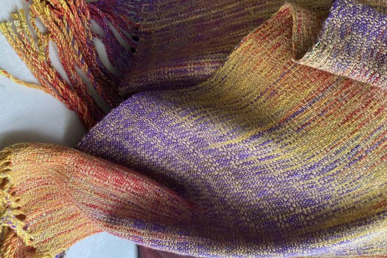 The image presents a close-up, textured view of a handwoven textile, likely a scarf or shawl, resting on a plain white surface. The fabric is richly colored and intricately woven, displaying a complex interplay of hues and patterns.