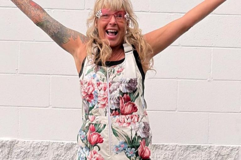 The image features a woman standing against a white brick wall with her arms raised in a celebratory gesture. She has blonde, curly hair with bangs and is wearing large, pink, novelty sunglasses with decorative details on the sides. Her mouth is open in a joyful expression. She is wearing a sleeveless, white outfit adorned with a vibrant floral print featuring large pink, red, and purple flowers, along with smaller blue flowers and green leaves. 