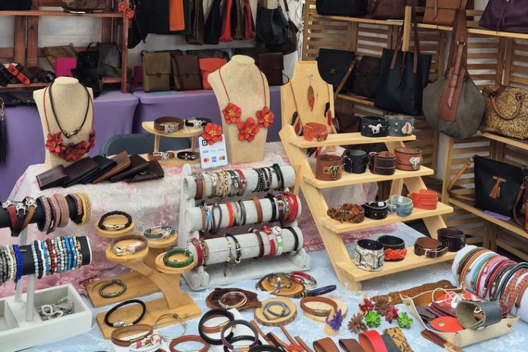 The image displays a vibrant market stall showcasing a variety of handmade leather goods and jewelry. The stall is arranged with multiple levels of wooden shelving and tables, creating an abundant and organized display.