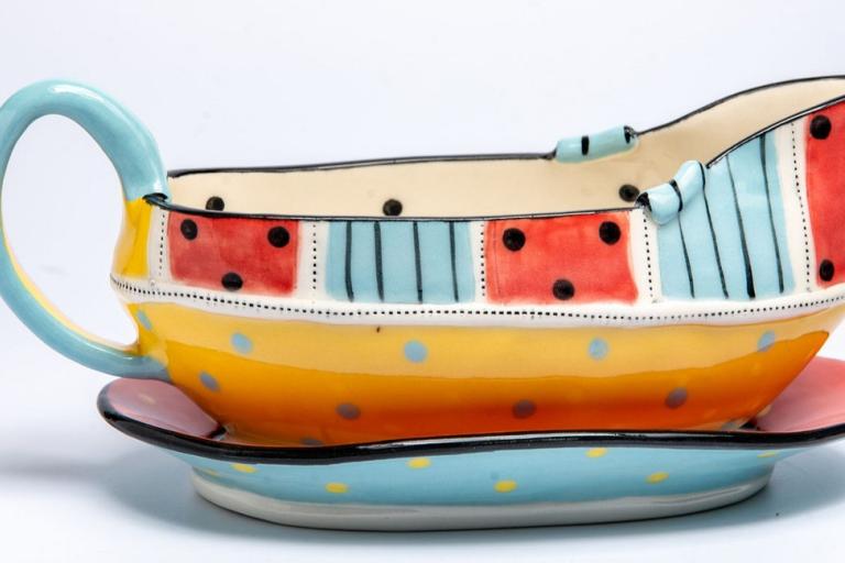 This is a studio shot of a decorative gravy boat and its accompanying saucer, presented against a clean white background. The gravy boat is creatively designed with a whimsical, almost childlike aesthetic. Its shape is elongated with a pouring spout at one end and a substantial handle arching from the opposite side.