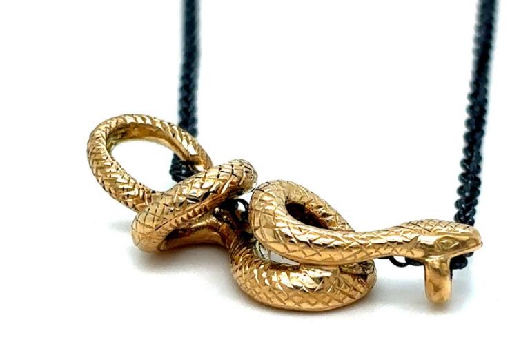 The image displays a close-up, eye-level shot of a gold necklace featuring a coiled snake pendant against a stark white background. The snake pendant is meticulously crafted, with its body intricately coiled and detailed to mimic the scales of a real snake. The head of the snake is depicted with a subtle curve, and the mouth is slightly open, with a small protrusion resembling a forked tongue or a decorative element. The snake is rendered in a lustrous gold color, catching the light and highlighting the tex