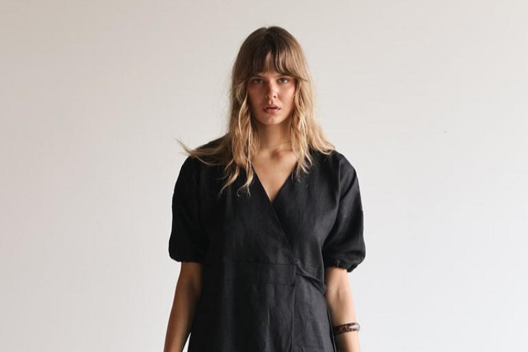 The image features a medium shot of a young woman with blonde hair and bangs, looking directly at the camera. She has a neutral expression, with her mouth slightly parted. Her hair is layered and falls around her shoulders. She is wearing a dark, possibly black, wrap dress with a V-neck and short, puffed sleeves. The dress appears to be made of a textured fabric, likely linen or a similar natural material, given its slightly rumpled appearance.