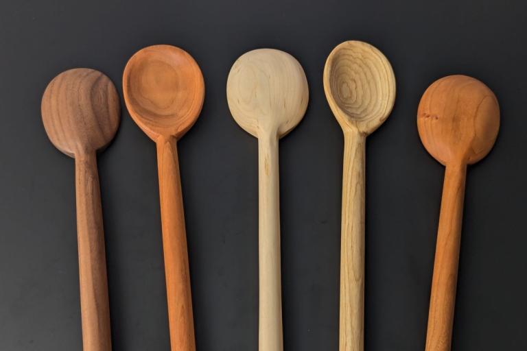 A top-down shot displays five wooden spoons arranged in a horizontal line against a dark, charcoal-colored background. The spoons are of varying sizes and wood types, creating a spectrum of natural brown and tan tones. From left to right, the first spoon is made of dark, reddish-brown wood, possibly walnut, with a smooth, rounded bowl and a long, flat handle. The second spoon is a lighter, orangey-brown, possibly cherry wood, with a similar shape to the first. The third spoon is the lightest, a pale blonde 