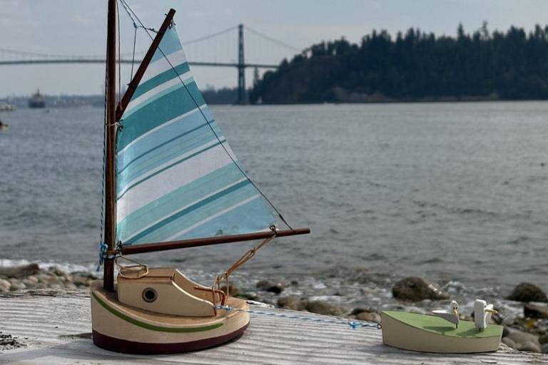 In the foreground of the image, two miniature toy boats are positioned on a weathered wooden surface that appears to be a pier or dock. The larger boat, on the left, is a sailboat with a predominantly light tan hull. It features a dark reddish-brown base and a thin green stripe just above it. The cabin area of the sailboat is a lighter tan color with a circular dark detail on its side. Its mast is made of wood, and the sail is the most striking element, displaying a pattern of alternating stripes in various