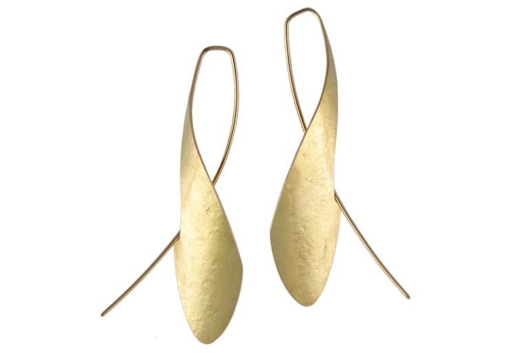 The image displays a pair of abstract, elongated drop earrings, likely made of brushed or hammered gold, against a stark white background. Each earring features a thin, delicate gold wire that forms a hook for pierced ears. From this hook, a larger, stylized pendant hangs. This pendant has a distinctive organic, teardrop or leaf-like shape that is slightly twisted or curved. The surface of the pendant is not smooth and polished but exhibits a textured, matte finish that suggests it has been brushed, hammere
