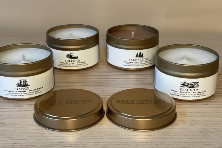 The image displays four lit candles in gold-toned metal containers arranged on a light-colored, wood-grain surface. The candles are positioned in a semi-circular formation with two candles in the front row and two slightly behind them. In front of the candles, two matching gold-toned lids are placed horizontally, with one slightly overlapping the other. Each candle container has a white label wrapped around its base. The labels feature text and a small illustration. The flame of each candle is visible and a