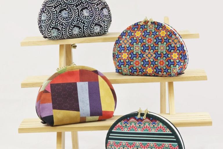 The image showcases a collection of four rounded, clutch-style purses arranged on a light-colored, tiered wooden shelf. The background is a plain, neutral off-white, suggesting a studio or product display setting. The lighting is even and bright, highlighting the textures and colors of the purses without harsh shadows.