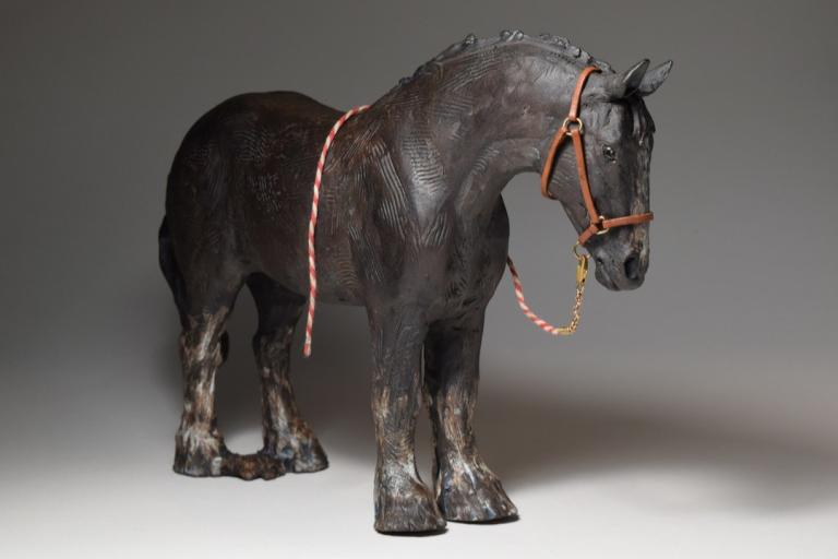 This is a close-up studio photograph of a dark, textured sculpture of a draft horse. The sculpture is dark brown, almost black, with visible tool marks and indentations across its body, suggesting a rough, earthy texture. The horse is depicted standing, with its head slightly bowed and turned to its right, facing towards the viewer. It wears a tan leather halter with brass-colored buckles and rings. A thin, red and white striped rope lead is attached to the halter and trails down and to the left, then loops