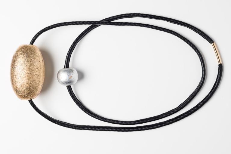 The image displays a piece of jewelry, likely a necklace or a bracelet, against a stark white background. The jewelry consists of a thick, black cord that is twisted into an oval shape. There are three distinct embellishments on the cord. On the left side, a large, oblong bead with a textured, golden surface is prominently featured. To its right, a smaller, spherical bead with a distressed, metallic silver finish is positioned. Towards the right side of the oval, the black cord is joined by a thin, cylindri