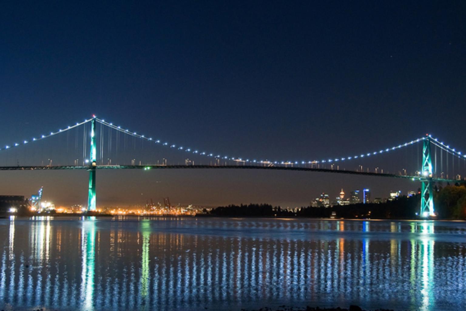 The Lions Gate Bridge seen at night with lights along the suspension cables. The water below reflect the bridges lights. 
