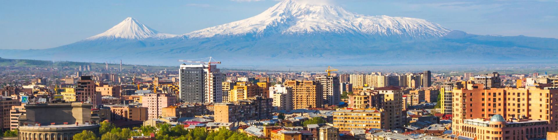 The image depicts a city with a backdrop of Mount Ararat, towering at 5,137 meters. The mountain is snow-capped and features two major volcanic cones. This view is from Yerevan, Armenia, set against a landscape that includes buildings, trees, and a cloudy sky. The scene combines urban elements with the natural beauty of the mountain and surrounding nature.