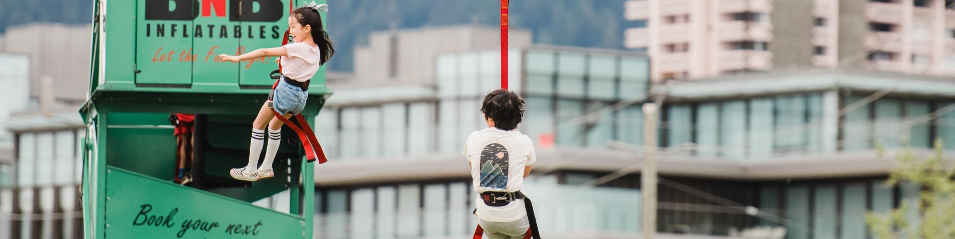 Two children on a rope swing in front of a building, enjoying a zipline ride together.
