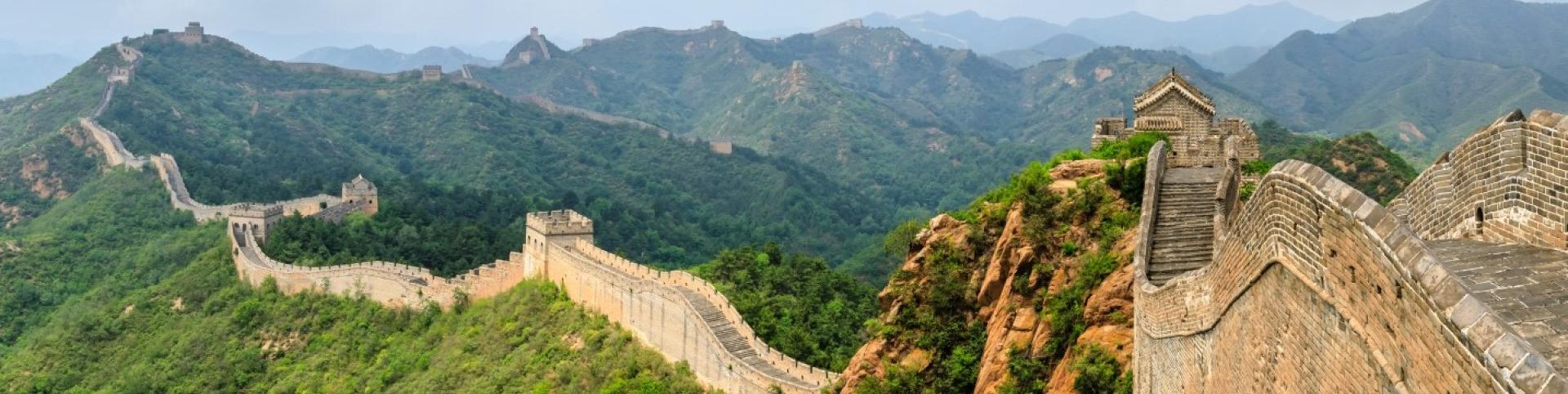 The image is of a stone wall on a hill, part of the Great Wall of China. The setting is outdoors with clouds and sky in the background, surrounded by a landscape with trees and mountains.