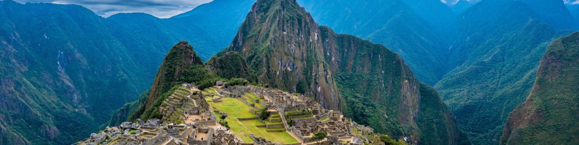 The image depicts a valley between mountains with Machu Picchu in the background. The setting is in Peru, showcasing a beautiful highland landscape with clouds, sky, and nature elements like grass and mountains.