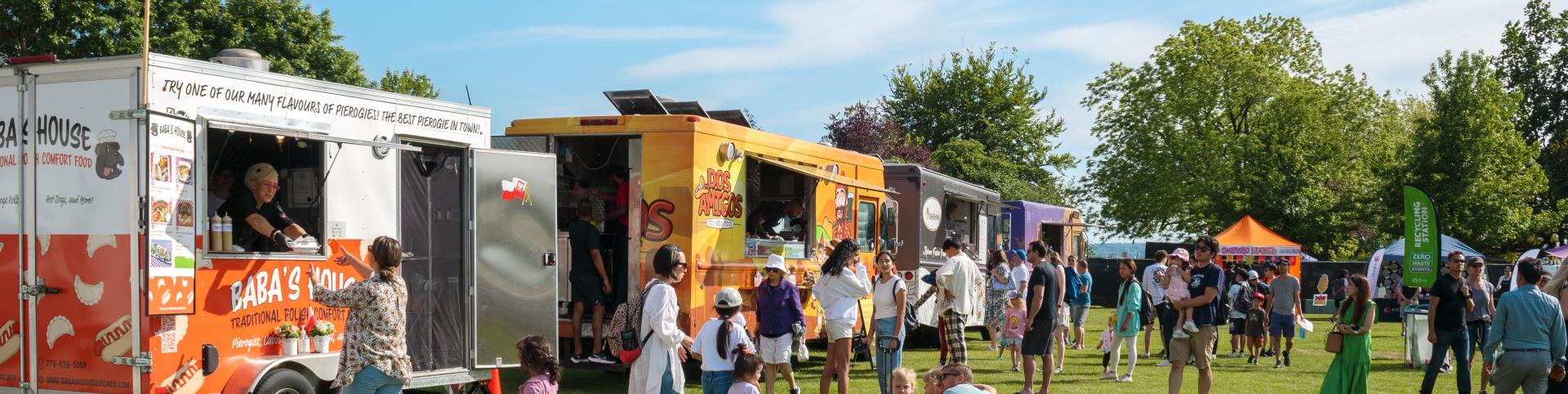 A group of people is gathered around a food truck, which is parked in an outdoor setting. The surroundings include grass and trees, with a clear sky overhead. The scene depicts a lively public space where individuals are likely enjoying the food being served from the mobile vendor.