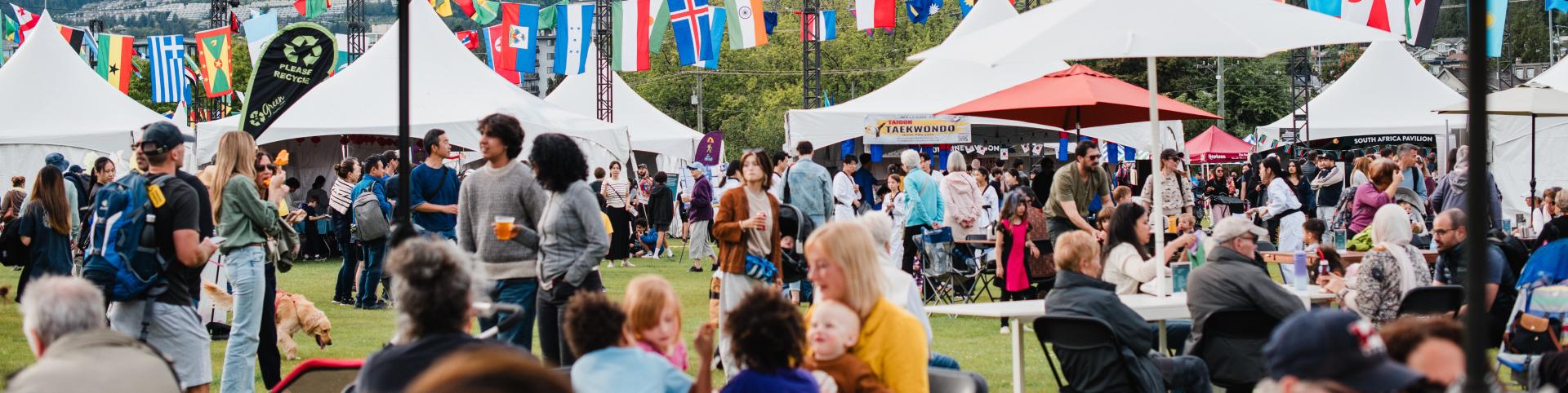 The image depicts a bustling crowd of people gathered at an outdoor event, likely a festival. Attendees are dressed in various clothing styles and are enjoying the activities under a canopy and tents. The setting features lush grass and nearby trees, with mountains visible in the background, contributing to the vibrant atmosphere of the occasion.